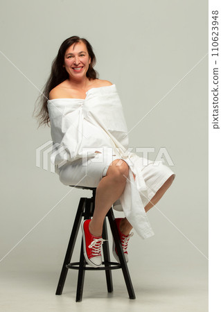A beautiful middle-aged woman wrapped in a white sheet poses on a studio chair. 110623948