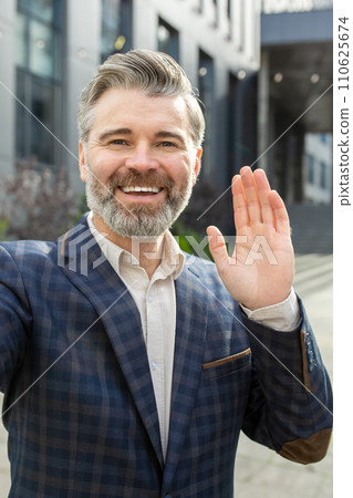 Smiling mature businessman in a stylish suit waving hello, standing outdoors by an office building, exuding leadership. 110625674