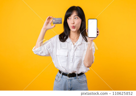 Portrait asian young woman wearing white shirt and denim jean with happy smile holding credit card to paying shopping online on smartphone isolated on yellow background. shopping online concept. Portrait asian young woman wearing white shirt and denim jean with happy smile holding credit card to paying shopping online on smartphone isolated on yellow background. shopping online concept. 110625852