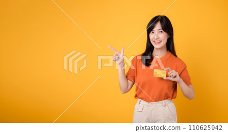 Portrait young beautiful 30s asian woman happy smile dressed in orange shirt showing credit card and point finger to free space against yellow background. Pay and purchase shopping payment concept. Portrait young beautiful 30s asian woman happy smile dressed in orange shirt showing credit card and point finger to free space against yellow background. Pay and purchase shopping payment concept. 110625942