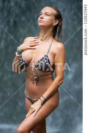 Beautiful young woman relaxes under a waterfall. Sexy girl in a bikini posing near a waterfall in the tropics. A traveler bathes under streams of water flowing from the mountains. 110625943