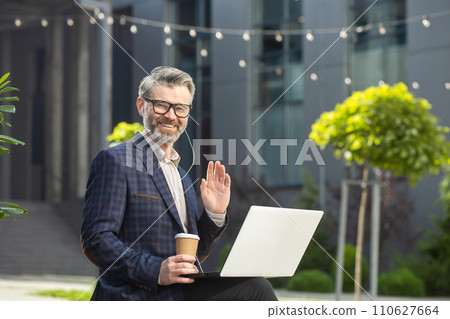 Friendly senior executive in business attire waving and working on a laptop outside a modern office building with a coffee cup. Friendly senior executive in business attire waving and working on a laptop outside a modern office building with a coffee cup. 110627664