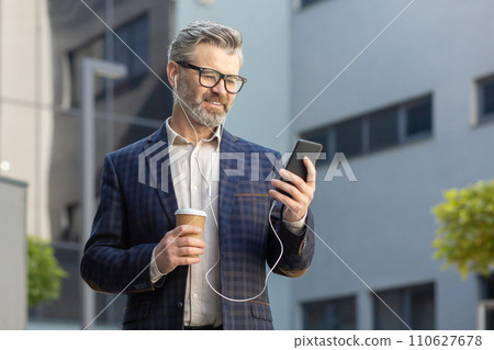Mature male executive in a suit with coffee cup texting on smartphone by a modern office building. 110627678