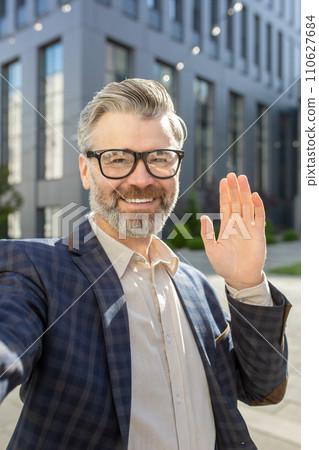 Cheerful senior male executive with glasses greeting with a hand wave, standing outside a modern office building in a suit. 110627684
