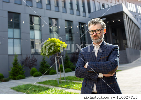 Mature executive with a serious expression standing confidently outside his workplace, arms crossed, embodying leadership. 110627715