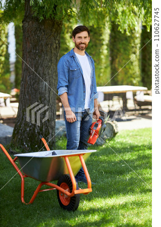 Smiling caucasian craftsman holding electric chainsaw, having break after work in garden. Front view of dark haired worker standing with wheelbarrow and battery saw for building. Concept of gardening. Smiling caucasian craftsman holding electric chainsaw, having break after work in garden. Front view of dark haired worker standing with wheelbarrow and battery saw for building. Concept of gardening. 110627745