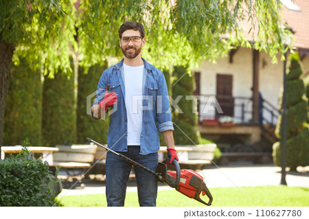Handsome gardener in gloves showing thumb up, while standing with electric hedge trimmer in garden. Portrait of happy brunet male worker making like gesture, while pruning plants. Concept of work. 110627780