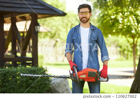 Confident gardener in protective glasses smiling, while posing with electric hedge trimmer outdoors. Portrait of happy handyman with bush cutter looking at camera, while working. Concept of lifestyle. 110627802