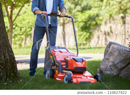 Unrecognizable male gardener in jeans using modern lawn mower on yard. Crop view of landscaper cutting overgrown grass with lawn trimmer, with blurred sunlit background. Concept of garden equipment. Unrecognizable male gardener in jeans using modern lawn mower on yard. Crop view of landscaper cutting overgrown grass with lawn trimmer, with blurred sunlit background. Concept of garden equipment. 110627822