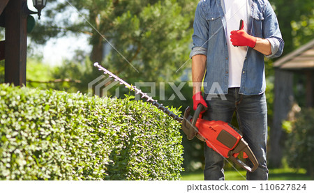 Anonymous muscular gardener in gloves showing thumb up, while standing with electric hedge trimmer at backyard. Crop view of male worker making like gesture, while pruning boxwood. Concept of work. 110627824