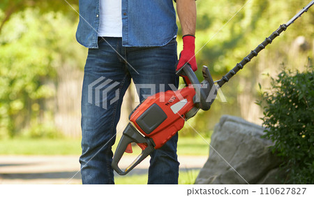 Unrecognizable muscular man in jeans standing with modern electric hedge trimmer outdoors. Crop view of strong male worker holding equipment for pruning, on blurred background. Concept of gardening. 110627827