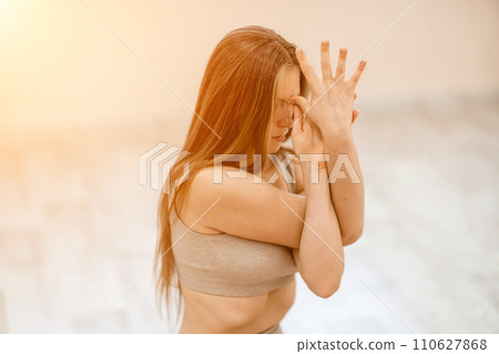 Young woman doing yoga in the gym. A girl with long hair and in a beige tracksuit stands in a cow pose on a pink carpet. A woman performs Gomukhasana. Young woman doing yoga in the gym. A girl with long hair and in a beige tracksuit stands in a cow pose on a pink carpet. A woman performs Gomukhasana. 110627868