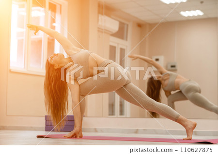 Young woman doing yoga in the gym. A girl with long hair and in a beige tracksuit stands in a parivritta trikonasana pose on a pink carpet. A woman performs parivritta trikonasana. 110627875