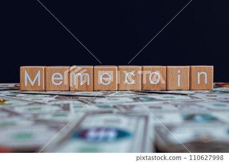 Wooden blocks with the text "Memecoin" on a black background and crypto banknotes scattered on the ground. 110627998