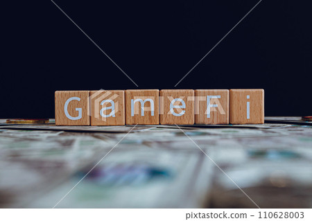 Wooden blocks with the text "GameFi" on a black background and crypto banknotes scattered on the ground. Wooden blocks with the text "GameFi" on a black background and crypto banknotes scattered on the ground. 110628003
