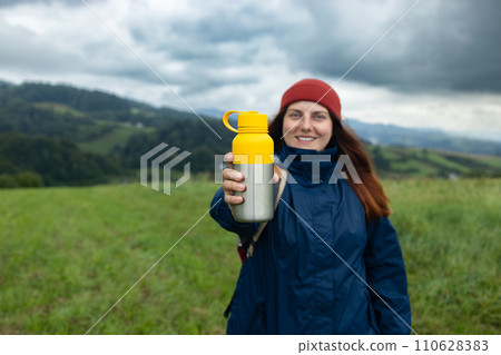 woman traveler drinks coffee with a view of the mountain landscape. A young tourist woman drinks a hot coffee drink and enjoys the scenery in the mountains. Trekking concept 110628383