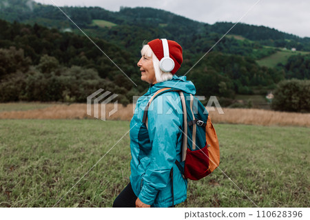 Happy active senior tourist woman listening to music in outdoor leisure trip admiring the landscape feeling inspired by the beauty of nature. 110628396