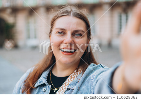 Smiling young woman taking a selfie while walking on a city street. Positive cheerful female student posing outdoors for sharing in social media app. Caucasian lady Smiling young woman taking a selfie while walking on a city street. Positive cheerful female student posing outdoors for sharing in social media app. Caucasian lady 110629362