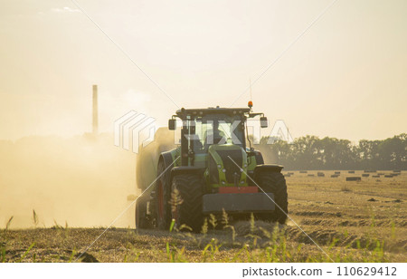 Combine harvester pressing straw from field into bales driving field on sunny summer evening. Field with bales of pressed wheat. Lots of dust on field. Agricultural agro-industrial harvesting works 110629412