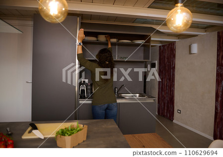 Rear view of brunette woman, a pretty housewife taking out plates from kitchen cabinet. People. Lifestyle. Domestic life Rear view of brunette woman, a pretty housewife taking out plates from kitchen cabinet. People. Lifestyle. Domestic life 110629694