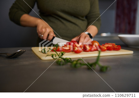 Close-up hands of female chef using kitchen knife, chopping tomatoes on wooden cutting board, standing at kitchen table 110629711