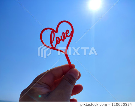 Person holding stick in shape red heart and word Love in hand on background of blue sky with shining sun on sunny summer day. Concept love relationship romance amour celebration St Valentines Day 110630554