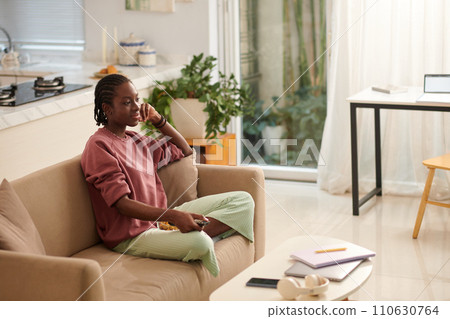 Bored teenage girl sitting on couch and watching true crime show 110630764