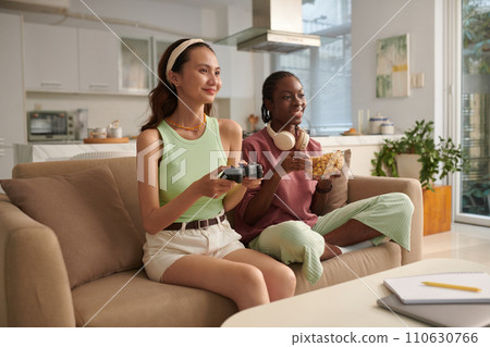 Smiling girl eating popcorn and watching her friend playing video game 110630766