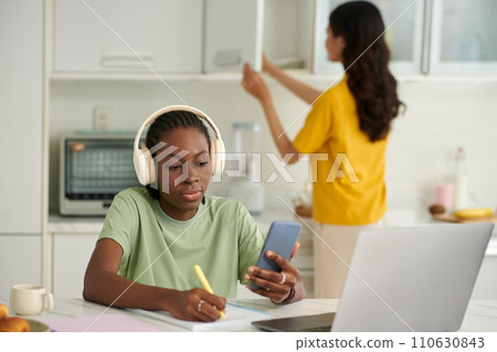 College student watching lection on smartphone and taking notes, when roommate cooking lunch College student watching lection on smartphone and taking notes, when roommate cooking lunch 110630843