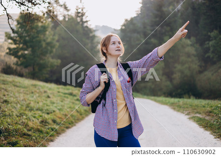 Blonde 30s girl pointing finger on forest background. Young female with backpack hiking on the road in nature. Beautiful woman walking in forest in sport clothes. Freedom and active lifestyle concept 110630902