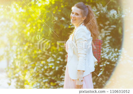 smiling trendy woman in pink dress and white jacket in city 110631052