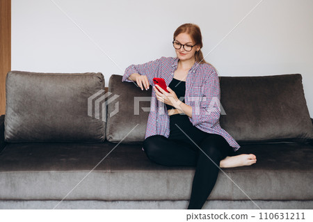 Young smiling woman holding smartphone, using cellphone modern technology, checking cell phone apps, texting, watching video, browsing internet for shopping. Girl sitting on sofa at home Young smiling woman holding smartphone, using cellphone modern technology, checking cell phone apps, texting, watching video, browsing internet for shopping. Girl sitting on sofa at home 110631211