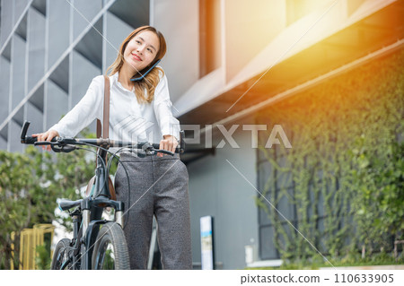 A businesswoman with her bike using a smartphone showcases the modern balance of work joy and technology. Her cheerful expression reflects the freedom to stay connected outdoors. A businesswoman with her bike using a smartphone showcases the modern balance of work joy and technology. Her cheerful expression reflects the freedom to stay connected outdoors. 110633905