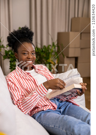 Black woman enjoying reading at home holding book while sitting on sofa. 110634420