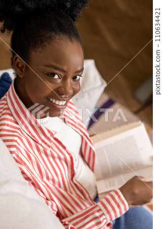 Black woman reading book while relaxing on couch in home interior. 110634421