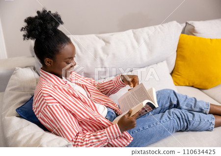 Smiling woman reading book while sitting on sofa at home. 110634425