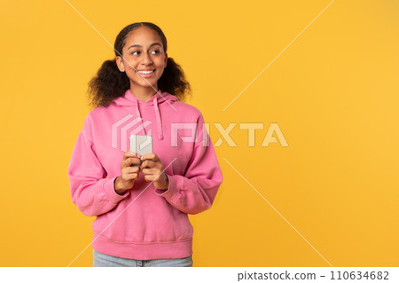 Cheerful black girl scrolling application on phone over yellow background 110634682