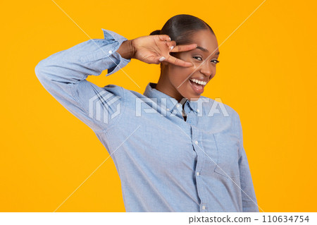 Black Young Woman Gesturing Victory Sign Near Face, Yellow Backdrop 110634754