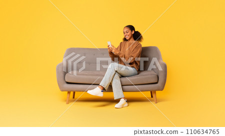 Black teen girl browsing on smartphone on couch, yellow backdrop Black teen girl browsing on smartphone on couch, yellow backdrop 110634765