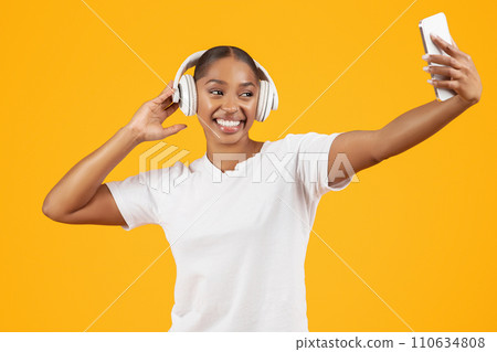 black lady making selfie listening music with earphones, yellow background 110634808