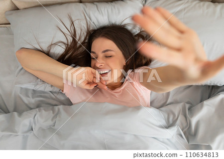 Joyful young lady stretching in bed with closed eyes and smile on her face after waking up in the morning, top view 110634813