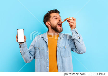 Guy eats pizza and shows mobile phone screen, blue backdrop 110634961