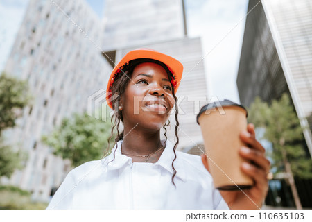 Happy black woman in helmet standing with coffee cup in daylight near buildings Happy black woman in helmet standing with coffee cup in daylight near buildings 110635103