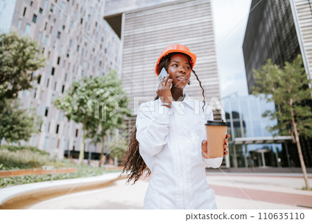 Happy black woman in helmet standing with coffee cup and speaking on smartphone 110635110
