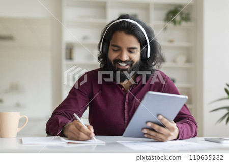 Indian man enjoying music on headphones while browsing tablet and taking notes 110635282