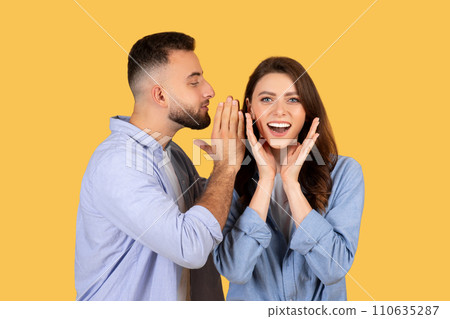 Man whispering to delighted woman with hands on cheeks on yellow background Man whispering to delighted woman with hands on cheeks on yellow background 110635287