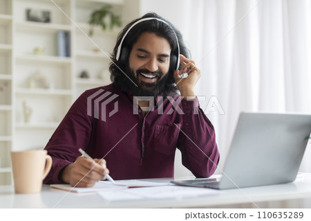 Distance Learning. Smiling Indian Man In Headphones Using Laptop And Taking Notes Distance Learning. Smiling Indian Man In Headphones Using Laptop And Taking Notes 110635289