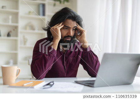 Concerned young indian man looking at laptop screen and holding his temples 110635339