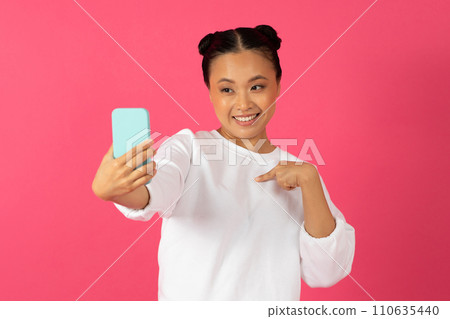 Happy young asian woman taking selfie on smartphone and pointing at herself Happy young asian woman taking selfie on smartphone and pointing at herself 110635440