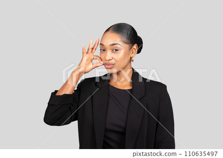 A cheerful young woman in a black suit makes a playful okay sign near her face 110635497